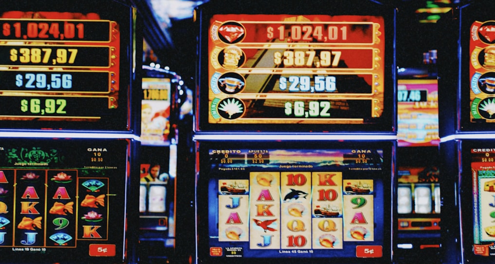 A row of colorful slot machines lit with bright neon lights, showcasing various graphical elements on the screens, indicating different games and jackpots. The atmosphere suggests a lively casino setting, bustling with excitement.
