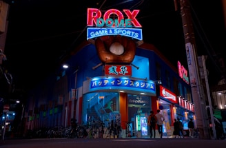 A vibrant, neon-lit facade of a gaming and sports establishment at night, featuring a large glove holding a baseball above the entrance. The building is adorned with colorful signs in both English and Japanese, with various advertisements and an illuminated sign that says 'ROX DOME'. Several people are walking towards the entrance, and bicycles are parked along the side.